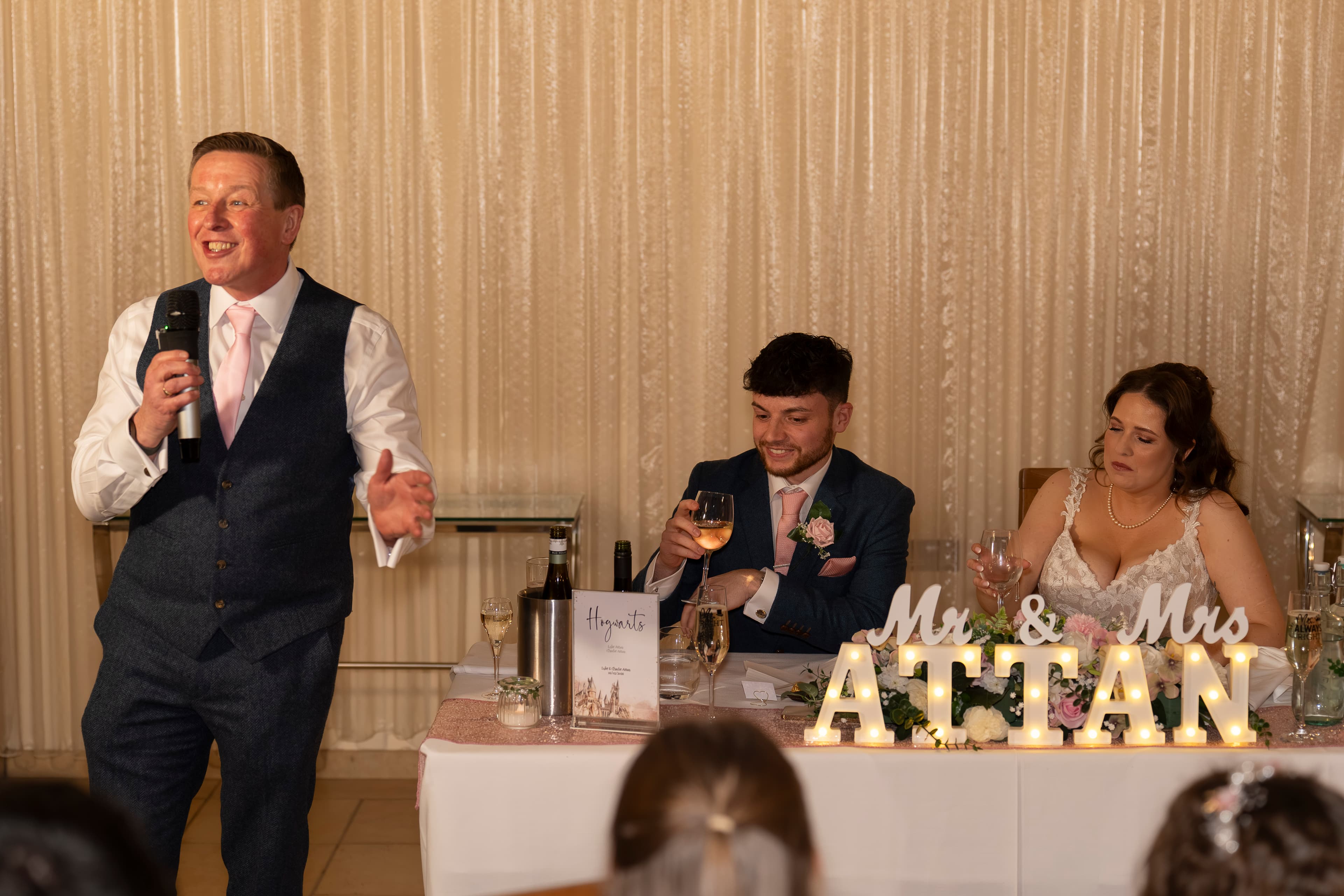 A wedding speaker holding a microphone beside the newlyweds at their reception.
